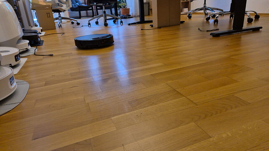 Roborock Saros 20 reinigt selbststaendig einen Teppich in einem Wohnzimmer. Im Hintergrund sitzt eine Familie entspannt auf dem Sofa. Lifestyle-Aufnahme zeigt alltagsnahen Einsatz des Saugroboters.